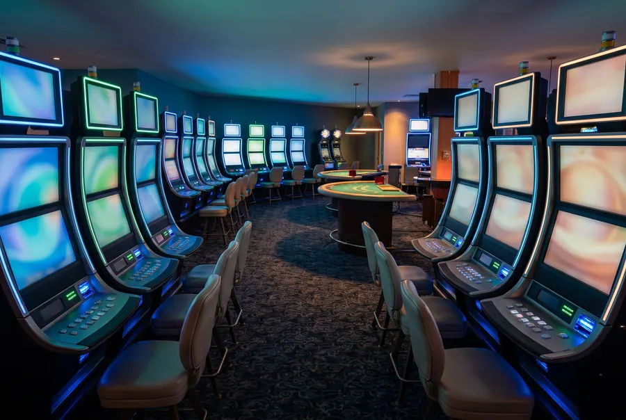Generated illustration: a dim gaming floor at night with curved rows of slot machines showing soft colour screens and an empty blackjack table under a lamp; not a photograph of the casino.